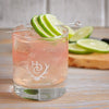 A Heritage Distilling Co. branded tumbler filled with a very light pink colored cocktail that is filled to the top with ice. A metal stir stick is place over top of the drink with three lime wedges over it. There are also more limes in the background.