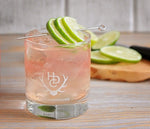 A Heritage Distilling Co. branded tumbler filled with a very light pink colored cocktail that is filled to the top with ice. A metal stir stick is place over top of the drink with three lime wedges over it. There are also more limes in the background.