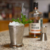 A metal mint julep cup is filled with the classic drink. HDC Elk Rider Bourbon and a double sided jigger are displayed in the backdrop of the photo.