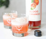 A bright orange colored cocktail with HDC Blood Orange Vodka in a salt-rimmed Heritage Distilling Co. - branded rocks glass set down on a white countertop.
