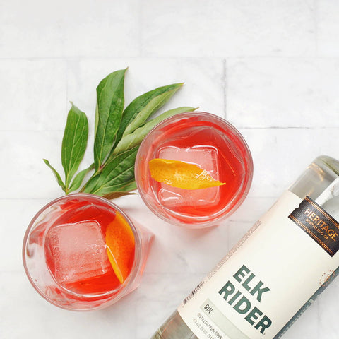 A bottle of HDC Elk Rider Gin is pictured with two cocktail glasses full of the Negroni cocktail with orange peel garnishes.
