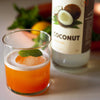 Vibrant orange colored cocktail poured over a single ice ball and garnished with a fresh mint leaf. HDC Coconut Vodka is featured behind the drink on a white table.