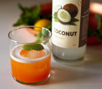 Vibrant orange colored cocktail poured over a single ice ball and garnished with a fresh mint leaf. HDC Coconut Vodka is featured behind the drink on a white table.