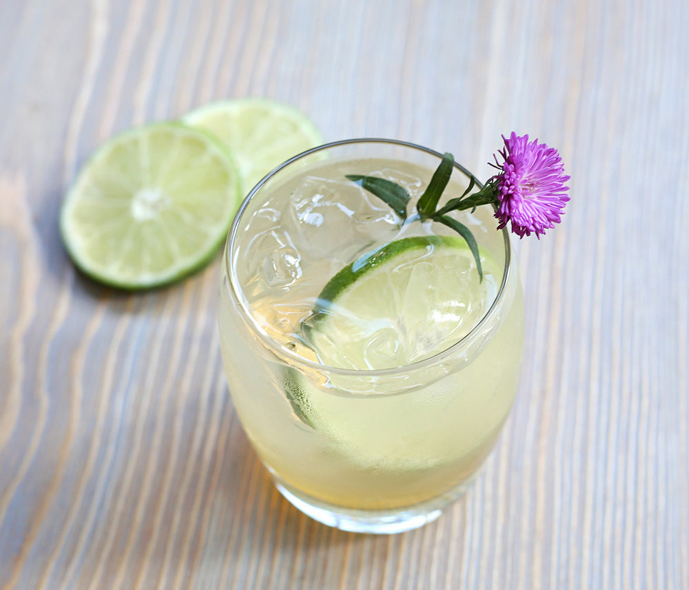 In a round tumbler, a light colored cocktail poured over ice with lime wheel garnishes.