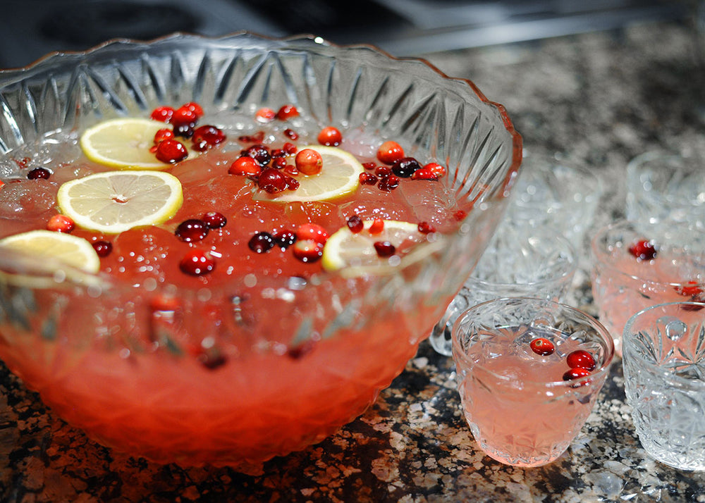 A crystal punch bowl full of a red vodka and bourbon based cocktail (using HDC Raspberry Vodka and BSB - Brown Sugar Bourbon). There are floating lemon wheels and fresh cranberries in the glasses of punch as well.