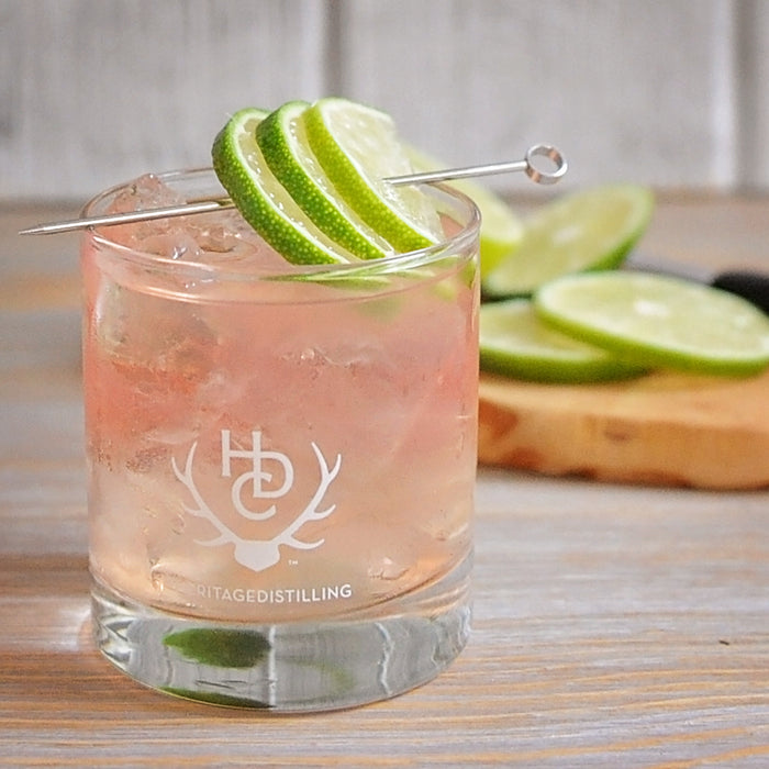 A Heritage Distilling Co. branded tumbler filled with a very light pink colored cocktail that is filled to the top with ice. A metal stir stick is place over top of the drink with three lime wedges over it. There are also more limes in the background.