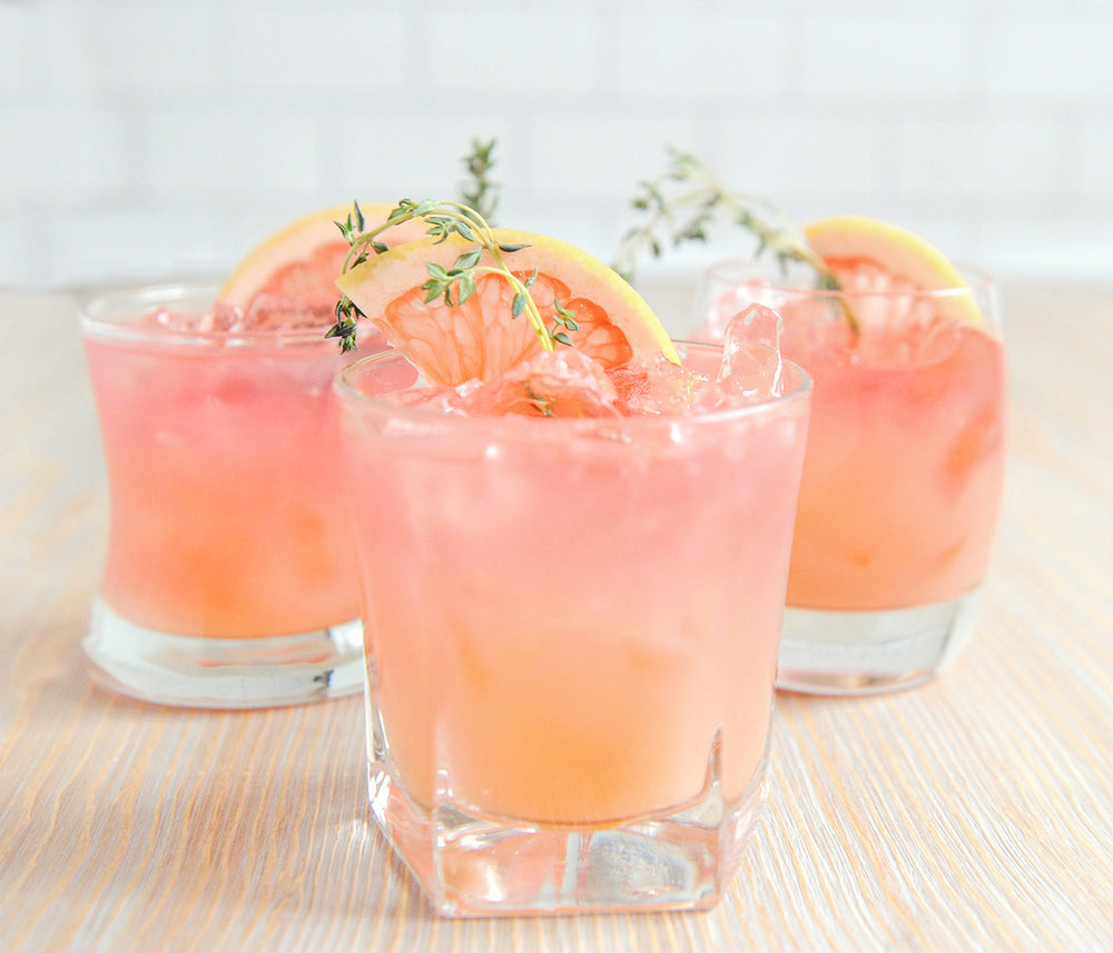 Three cocktail tumbler glasses full of ice with a light pink and orange drink garnished with grapefruit wedges.