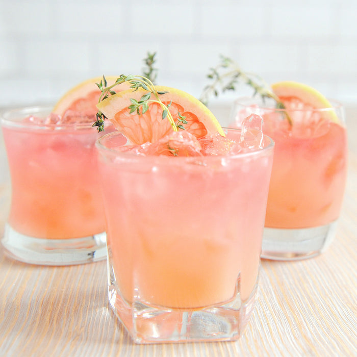 Three cocktail tumbler glasses full of ice with a light pink and orange drink garnished with grapefruit wedges.