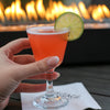 Red cocktail served up in a cocktail goblet with a lime wheel garnish. Enjoyed outside by a live flame fire pit.