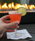 Red cocktail served up in a cocktail goblet with a lime wheel garnish. Enjoyed outside by a live flame fire pit.