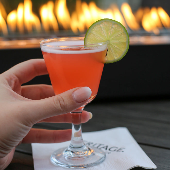 Red cocktail served up in a cocktail goblet with a lime wheel garnish. Enjoyed outside by a live flame fire pit.