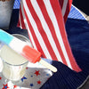 Red, white and blue popsicle balanced on top of a glass containing vodka lemonade. Both sitting on top of a star spangled napkin with an American flag in the background.