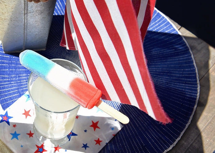 Red, white and blue popsicle balanced on top of a glass containing vodka lemonade. Both sitting on top of a star spangled napkin with an American flag in the background.