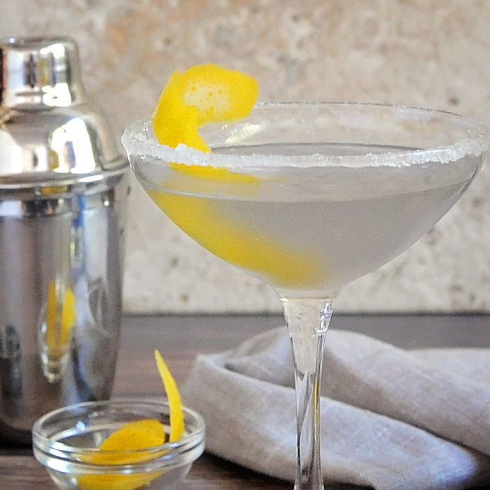 A sugar-rimmed cocktail glass with a vodka cocktail and a lemon peel garnish. A metal shaker is displayed next to the drink.