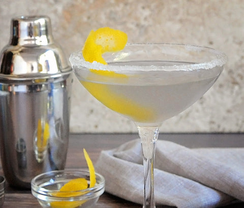A sugar-rimmed cocktail glass with a vodka cocktail and a lemon peel garnish. A metal shaker is displayed next to the drink.