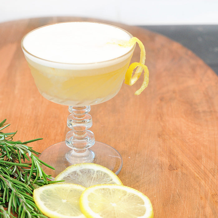 A cocktail goblet with the Rosemary Maple Whiskey Sour featuring HDC Elk Rider Bourbon and garnished with a lemon twist.