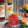 Bottles of HDC Blood Orange Vodka and HDC Citrus Vodka are shown behind this fruity cocktail. 