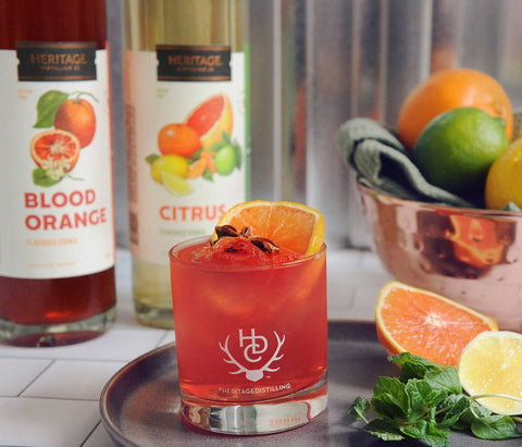 Bottles of HDC Blood Orange Vodka and HDC Citrus Vodka are shown behind this fruity cocktail. 