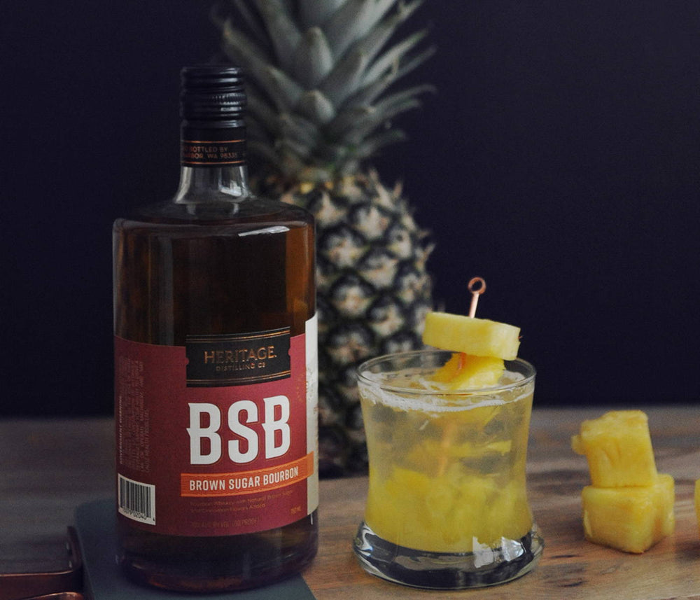 A 750ml bottle of BSB - Brown Sugar Bourbon, a whole pineapple, and a cocktail made with your choice of either BSB - Brown Sugar Bourbon or BSB 103. The garnishes are pineapple chunks.