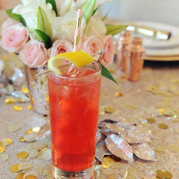 A three ingredient red cocktail with HDC Pomegranate vodka as its spirit base with a lemon twist garnish. The drink is placed on a table near a beautiful vase full of pink roses.