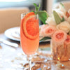 A pink and orange-colored cocktail in a champagne flute with blood orange slice and mint. The drink is photographed on a table with a bowl full of pink and white roses.