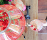 This recipe makes a shareable batch of Raspberry Thyme Lemonade (shown in a large serving container) with a glass full of the cocktail. It is garnished with fresh raspberries and thyme.