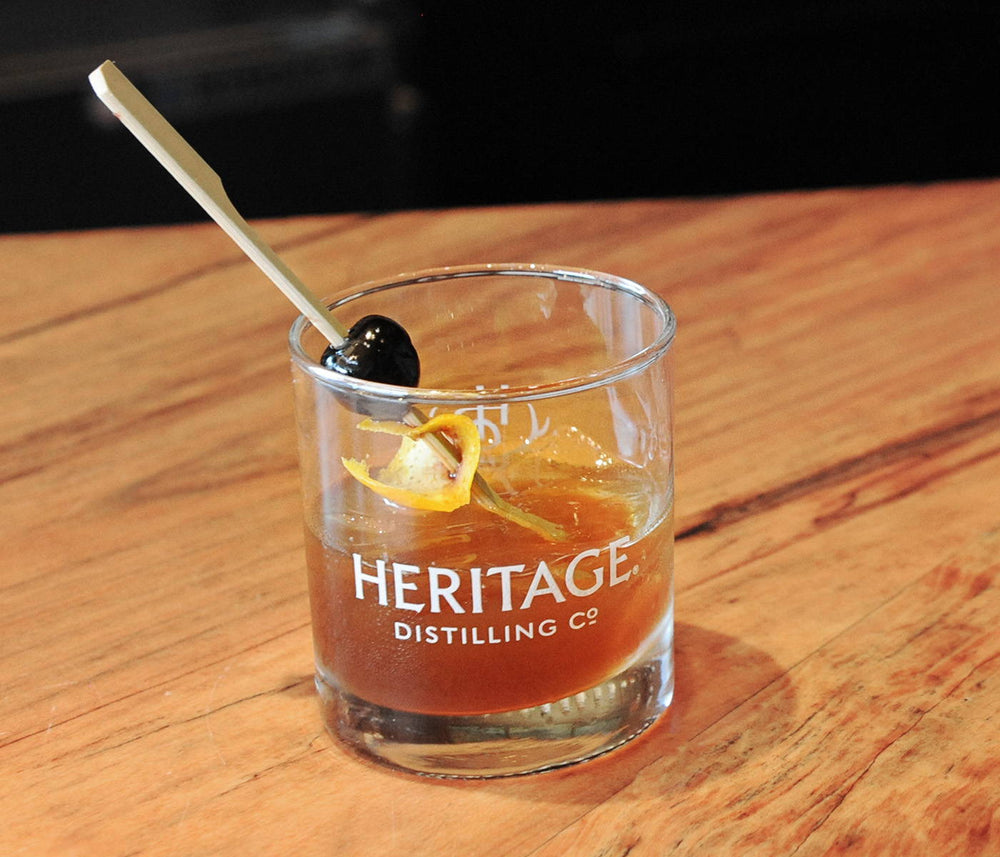 Heritage Distilling Co. branded rocks glass with a darker orange colored cocktail with only around 5 ice cubes in the glass with a stirring stick that has a Luxardo cherry and orange peel placed into the center of the drink
