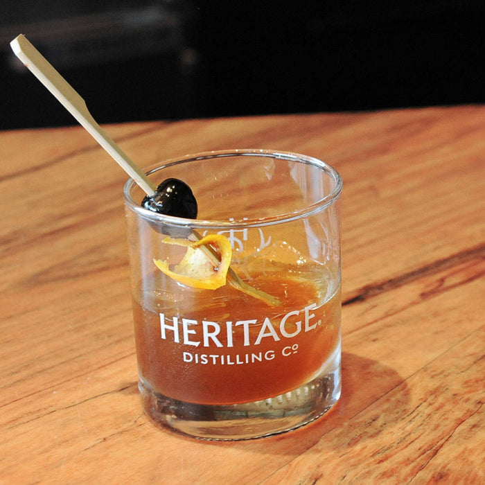 Heritage Distilling Co. branded rocks glass with a darker orange colored cocktail with only around 5 ice cubes in the glass with a stirring stick that has a Luxardo cherry and orange peel placed into the center of the drink