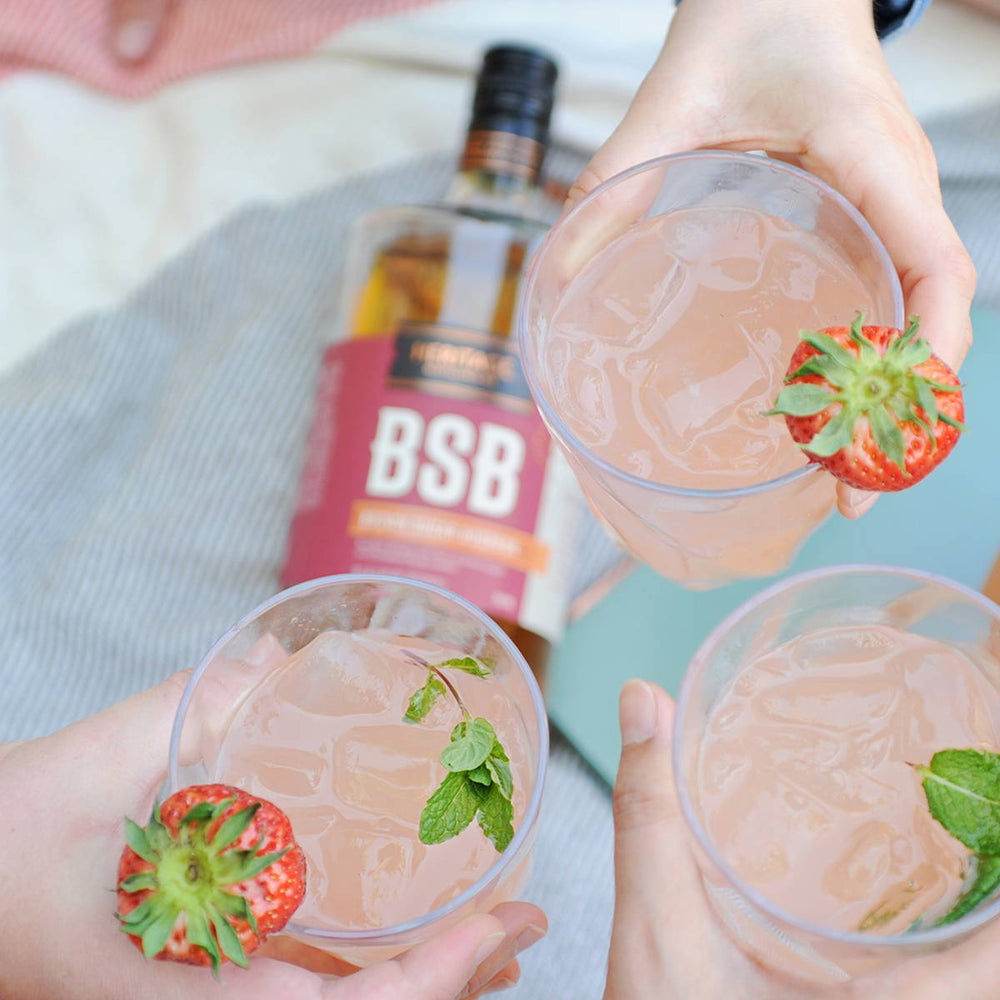 A full bottle of HDC BSB Brown Sugar Bourbon is in the background of three people's hands featuring their light pink cocktails served over ice and garnished with fresh strawberries