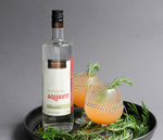 A bottle of HDC Bestemors Aquavit (a Scandinavian spirit) and two full glasses of the Aquavit Greyhound cocktail are on a tray.