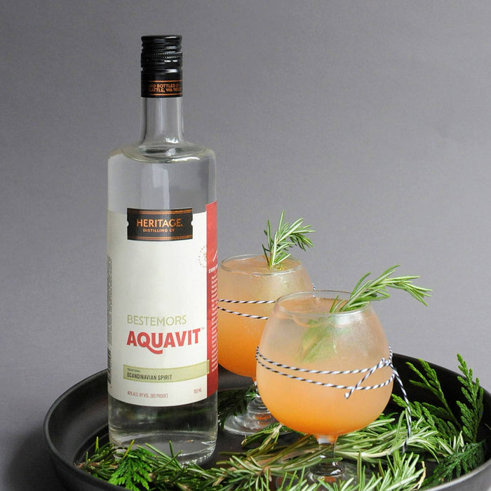 A bottle of HDC Bestemors Aquavit (a Scandinavian spirit) and two full glasses of the Aquavit Greyhound cocktail are on a tray.