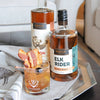One bottle of HDC Elk Rider Bourbon and one of HDC Bacon are displayed on a silver tray along with a cocktail with bacon slices and a Luxardo cherry as garnishes.