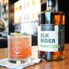 A bottle of HDC Elk Rider Rye is placed on top of a Heritage Distilling Co. Tasting Room wooden bar top. The full to the brim light orange cocktail, is garnished with a lemon twist.