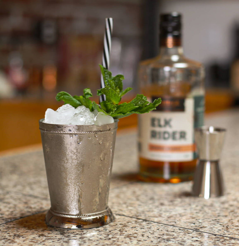 A metal mint julep cup is filled with the classic drink. HDC Elk Rider Bourbon and a double sided jigger are displayed in the backdrop of the photo.