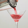 A shaker with a strainer contains a pinkish red cocktail being poured into a cold martini glass.