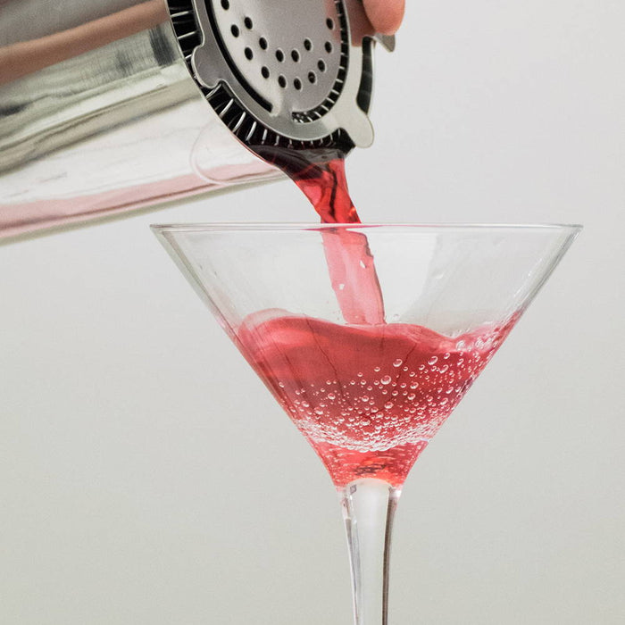 A shaker with a strainer contains a pinkish red cocktail being poured into a cold martini glass.