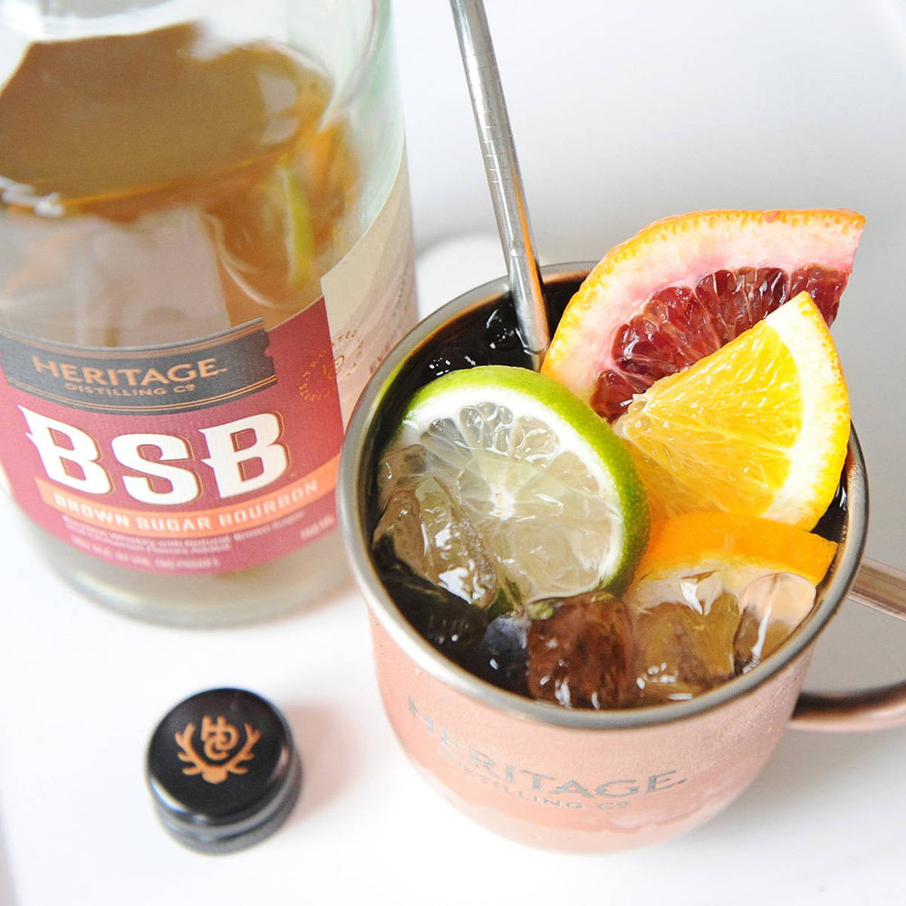 A bottle of BSB - Brown Sugar Bourbon and a BSB Citrus Mule in a copper mug filled with ice and fruit garnishes.