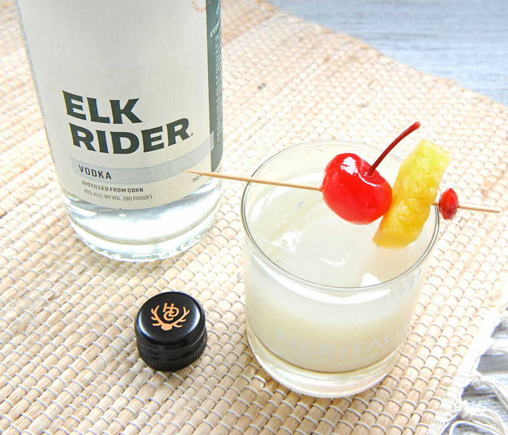 A bottle of HDC Elk Rider Vodka is displayed on a picnic blanket with a white cocktail poured over a rocks glass of ice with a cherry and pineapple garnish next to the spirits bottle.