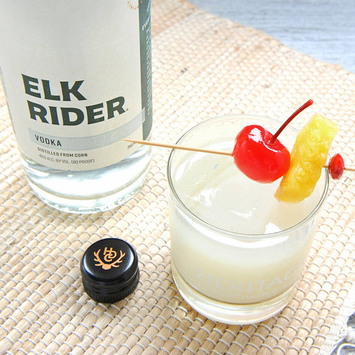 A bottle of HDC Elk Rider Vodka is displayed on a picnic blanket with a white cocktail poured over a rocks glass of ice with a cherry and pineapple garnish next to the spirits bottle.