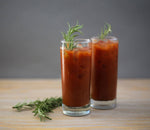 Two tall glasses of classic red-colored vodka Bloody Mary cocktail with generous fresh rosemary garnishes in the drinks and placed on a wooden table