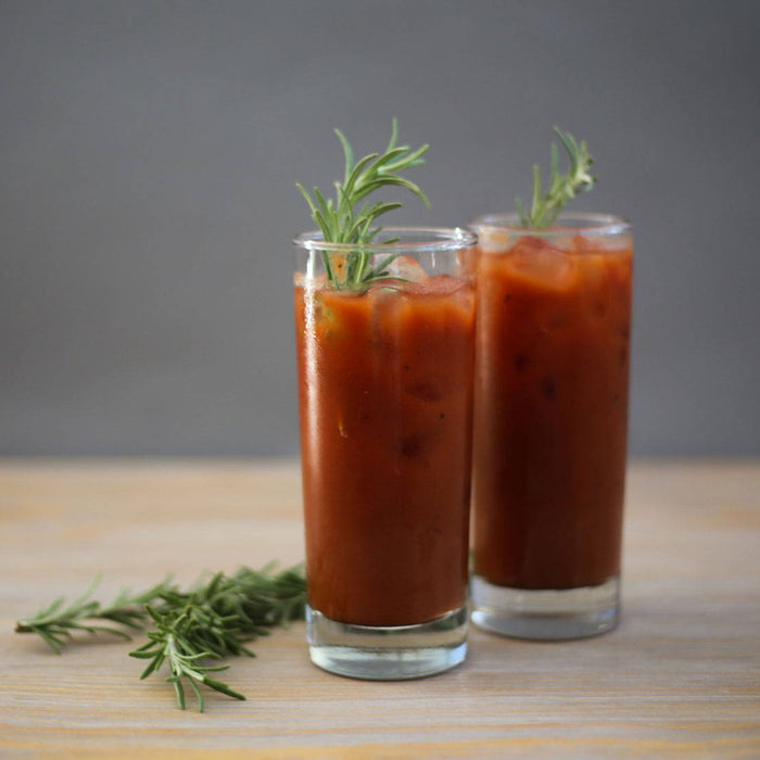 Two tall glasses of classic red-colored vodka Bloody Mary cocktail with generous fresh rosemary garnishes in the drinks and placed on a wooden table