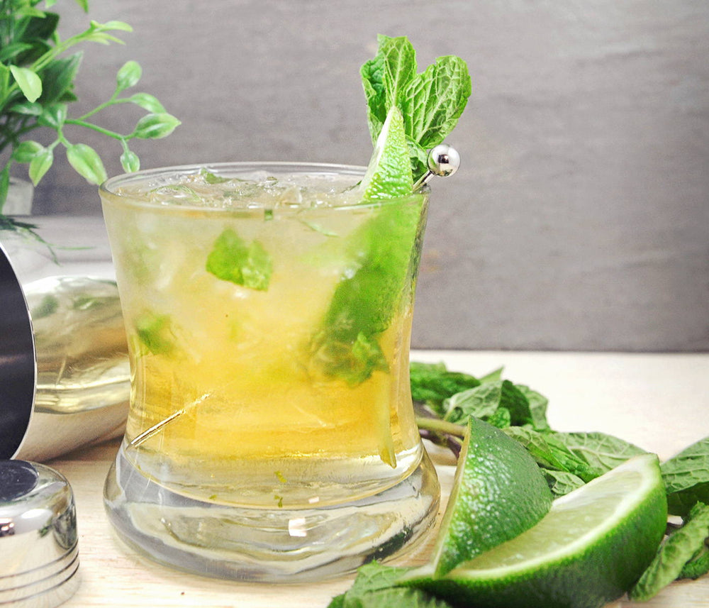 A whiskey cocktail poured into a glass of crushed ice. Lime wedges and mint sprigs are both in the drink and placed on the table top.