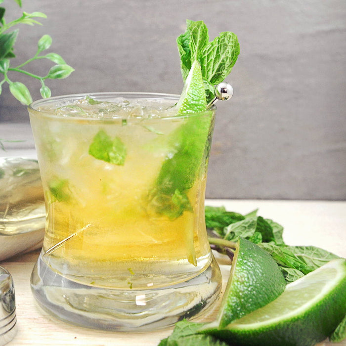 A whiskey cocktail poured into a glass of crushed ice. Lime wedges and mint sprigs are both in the drink and placed on the table top.