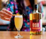A bottle of BSB - Brown Sugar Bourbon is displayed on a wooden countertop. bourbon cocktail is poured into a daisy cocktail glass with an orange peel garnish and Luxardo cherry on a stir stick.