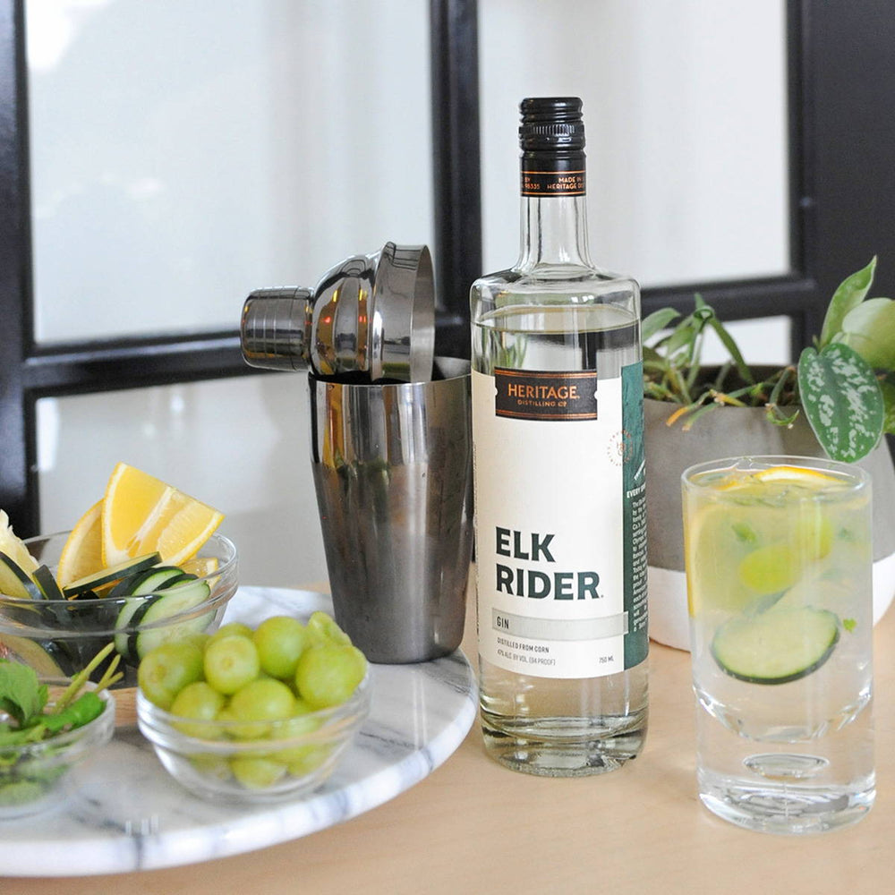 A 750ml bottle of Elk Rider Gin, a full cocktail, a metal shaker, and some garnish options (such as: grapes, lemons, and cucumbers).