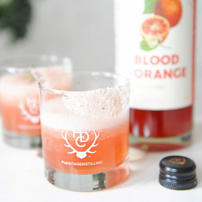 A bright orange colored cocktail with HDC Blood Orange Vodka in a salt-rimmed Heritage Distilling Co. - branded rocks glass set down on a white countertop.