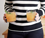 A person in a striped shirt is holding two of glasses of the Rosemary Ginger Cider that features BSB - Brown Sugar Bourbon.