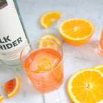 A bottle of HDC Elk Rider Gin is pictured next to a bright orange cocktail poured into a glass with one big single square-shaped ice cube. The cocktail is garnished with an orange peel and orange slices on the countertop for a fresh look.