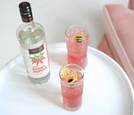 On a white circular table, there is a display bottle of HDC Sweet Ghost Pepper Vodka and two highball glasses filled with ice and a pink cocktail with lemon twists and a cherry as garnishes.