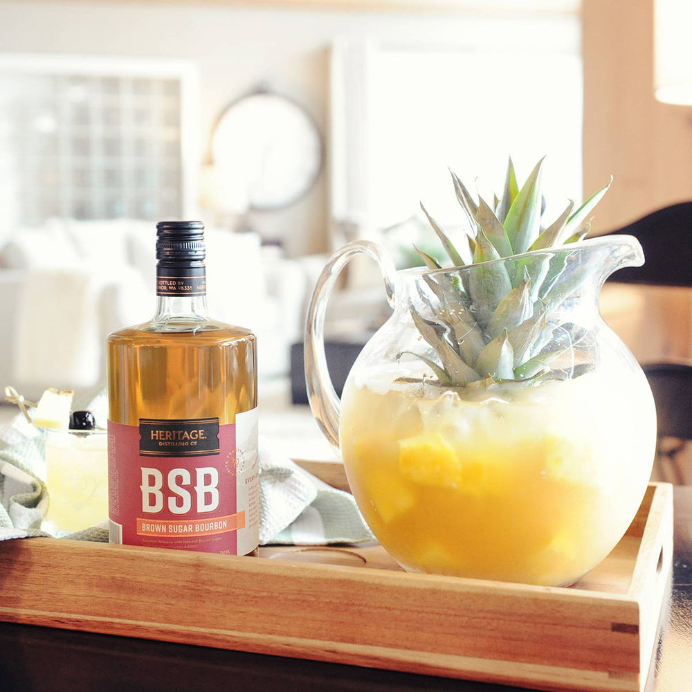 A bottle of BSB - Brown Sugar Bourbon with a pitcher full of this pineapple and bourbon cocktail with many pineapple chunks in the batch.